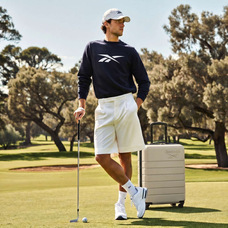 A man in golf attire stands on a course with a putter beside the beige Reebok Captain 2 Piece Luggage Set with spinner wheels. Trees and green grass fill the background.