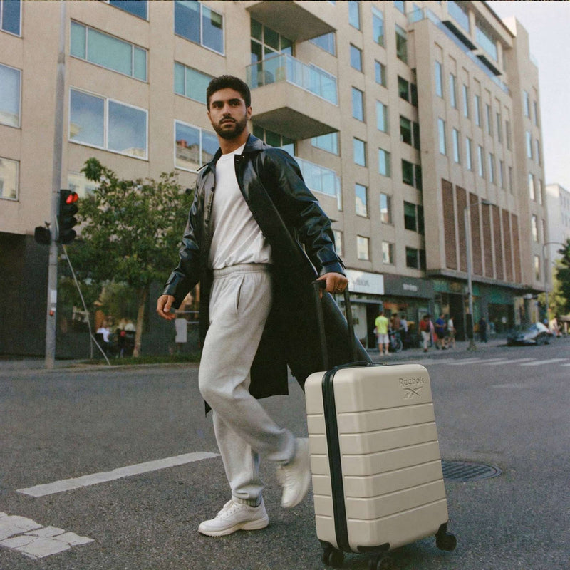 A man in a black leather coat and gray sweats crosses a city street, pulling the cream-colored Reebok Captain 2 Piece Luggage Set with spinner wheels. Modern buildings and pedestrians are visible in the background.