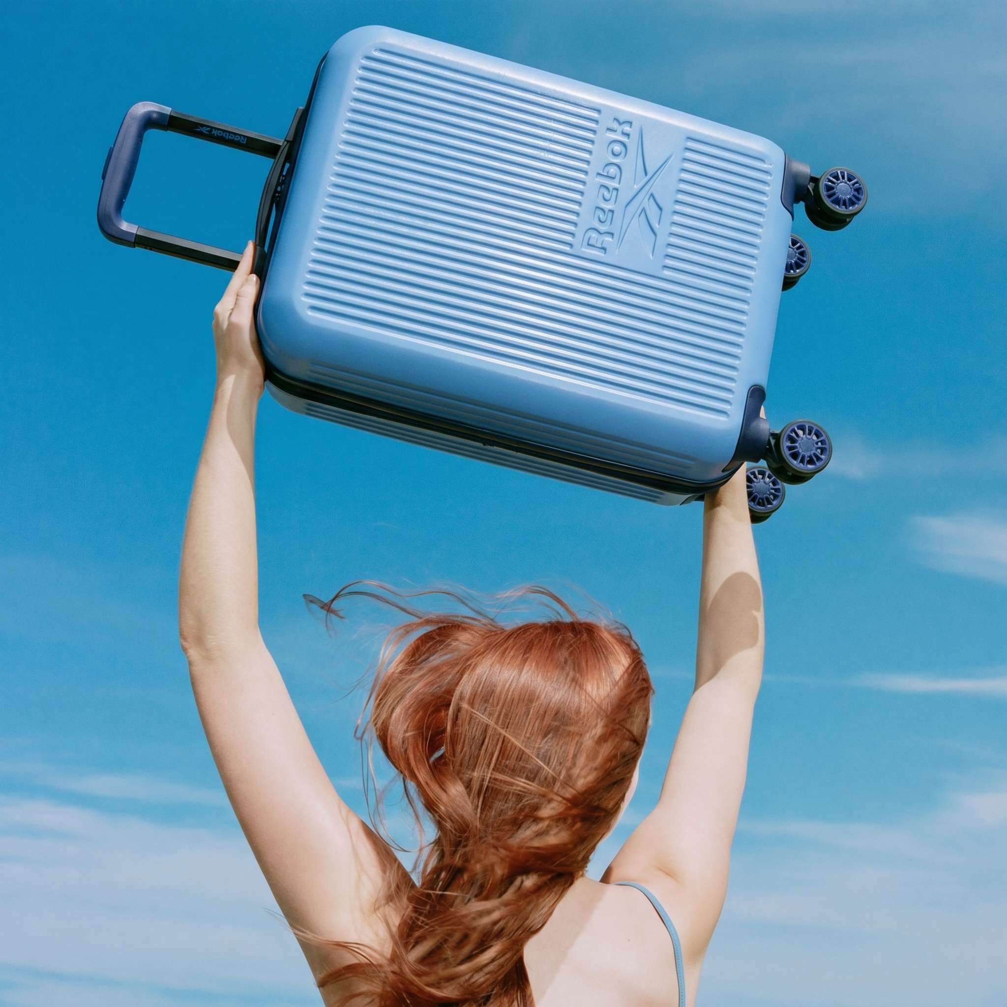 A person with long red hair lifts a Reebok Action Duo 2 Piece Luggage Set suitcase with spinner wheels overhead against a bright blue sky with wispy clouds.