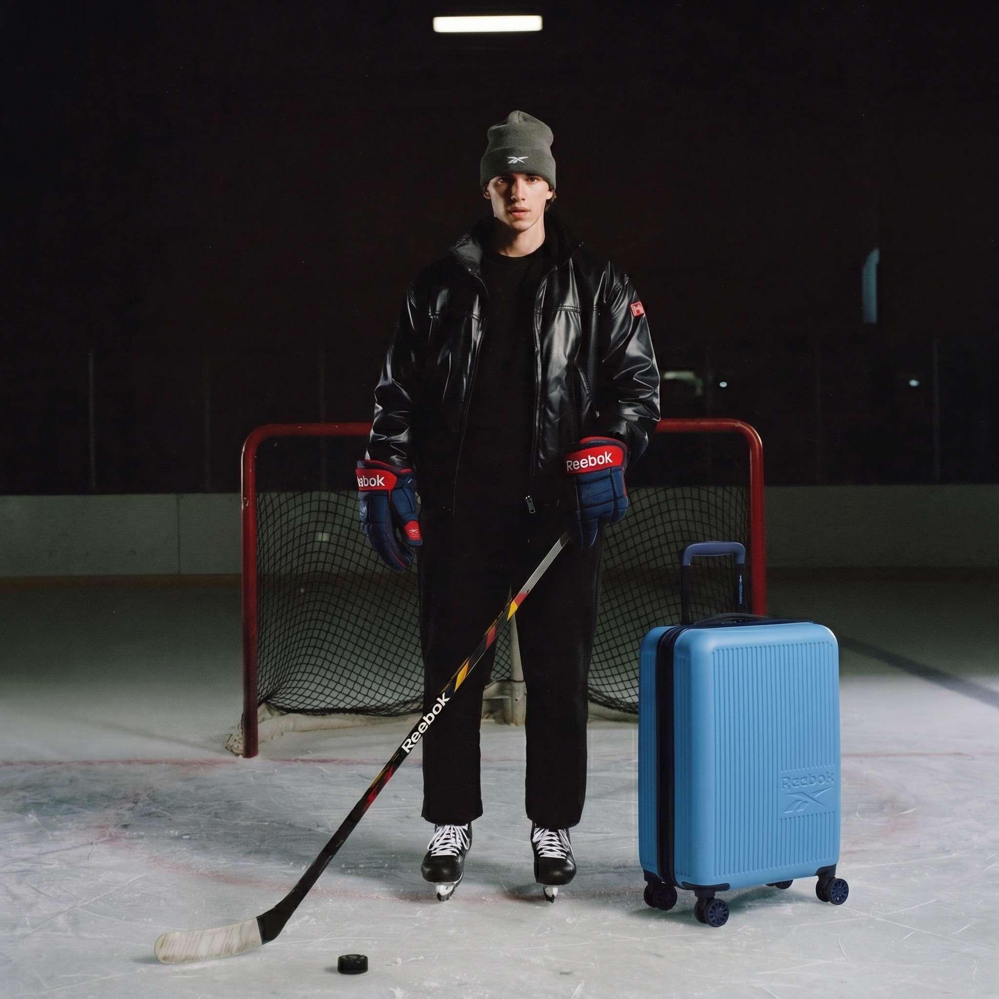 A hockey player, wearing Reebok gear, stands on the ice with a stick, puck, and the Action Duo 2 Piece Luggage Set by their side.