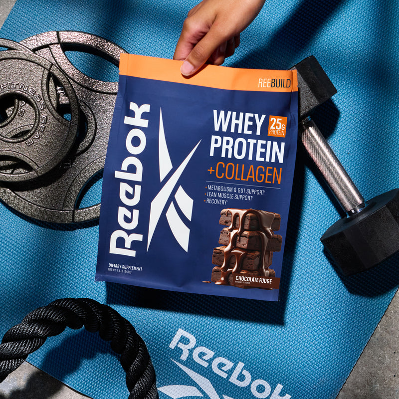 A hand holds a pouch of Reebok ReeBuild Whey Protein + Collagen over a blue gym mat with weight plates and a dumbbell.
