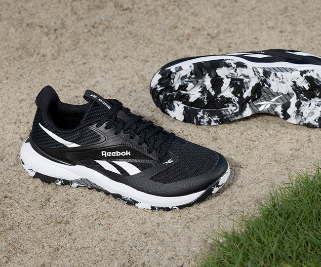 A pair of black Reebok athletic shoes with white accents and marbled black-and-white soles are displayed on sandy ground near a patch of green grass.