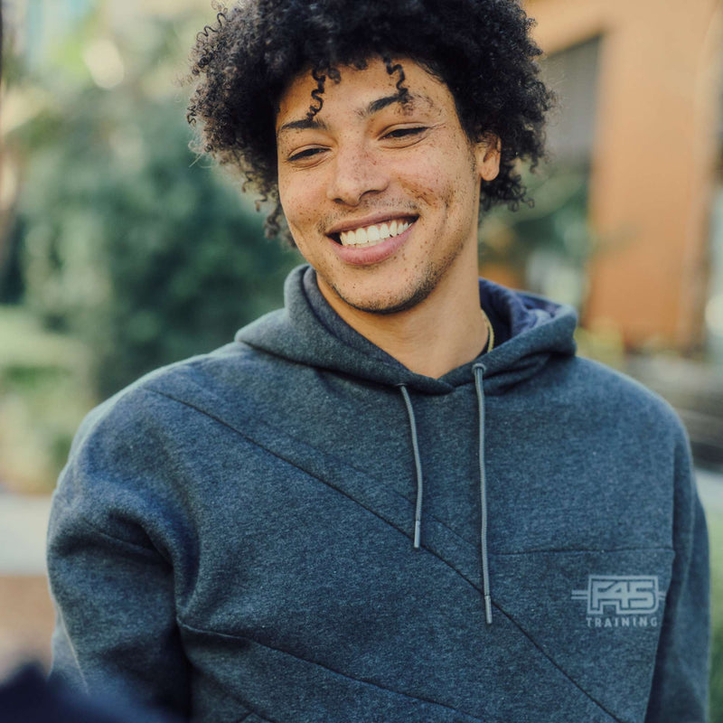 A young man with curly hair and freckles smiles while wearing the Men's Reebok x F45 Training Relaxed Hoodie outdoors, with greenery and a blurred building in the background.