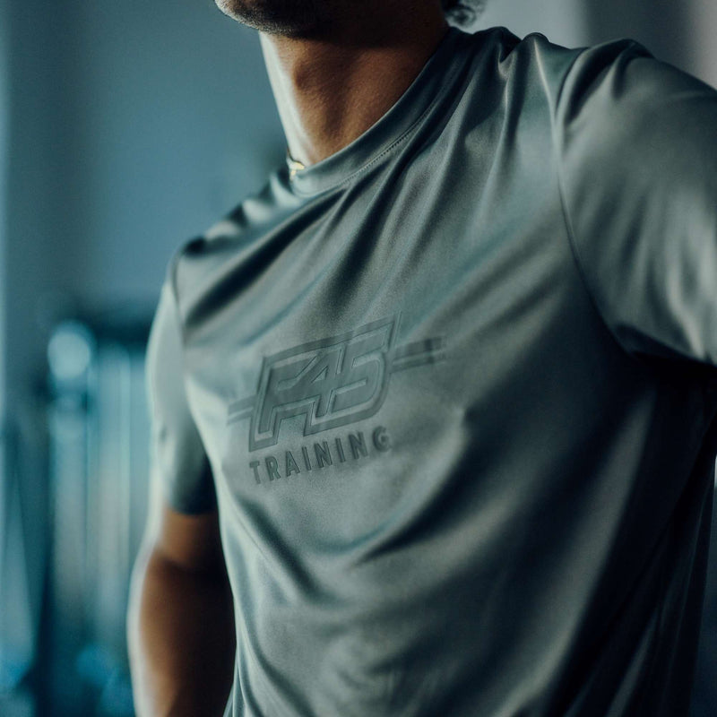 Close-up of a person wearing the Men's Reebok x F45 Training T-Shirt, featuring the Reebok brand logo prominently displayed on the chest. The gray athletic shirt stands out in focus, while a blurred background hints at an indoor gym or fitness environment.