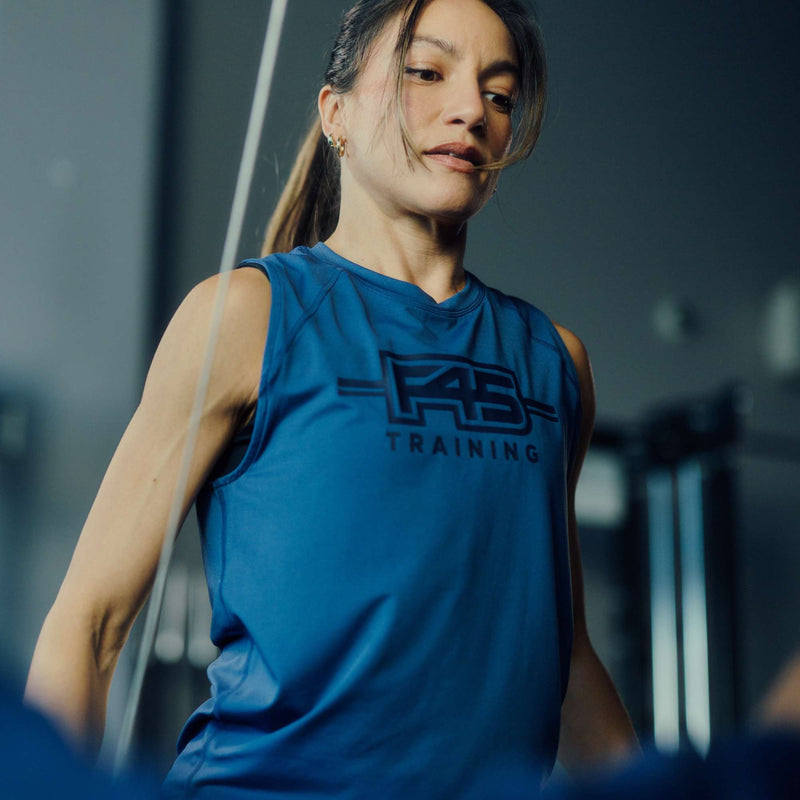 A woman wears the Women's Reebok x F45 Training Tank Top, focused as she exercises indoors. The blurred background shows gym equipment and dark tones, highlighting the Reebok branding and sweat-wicking design.