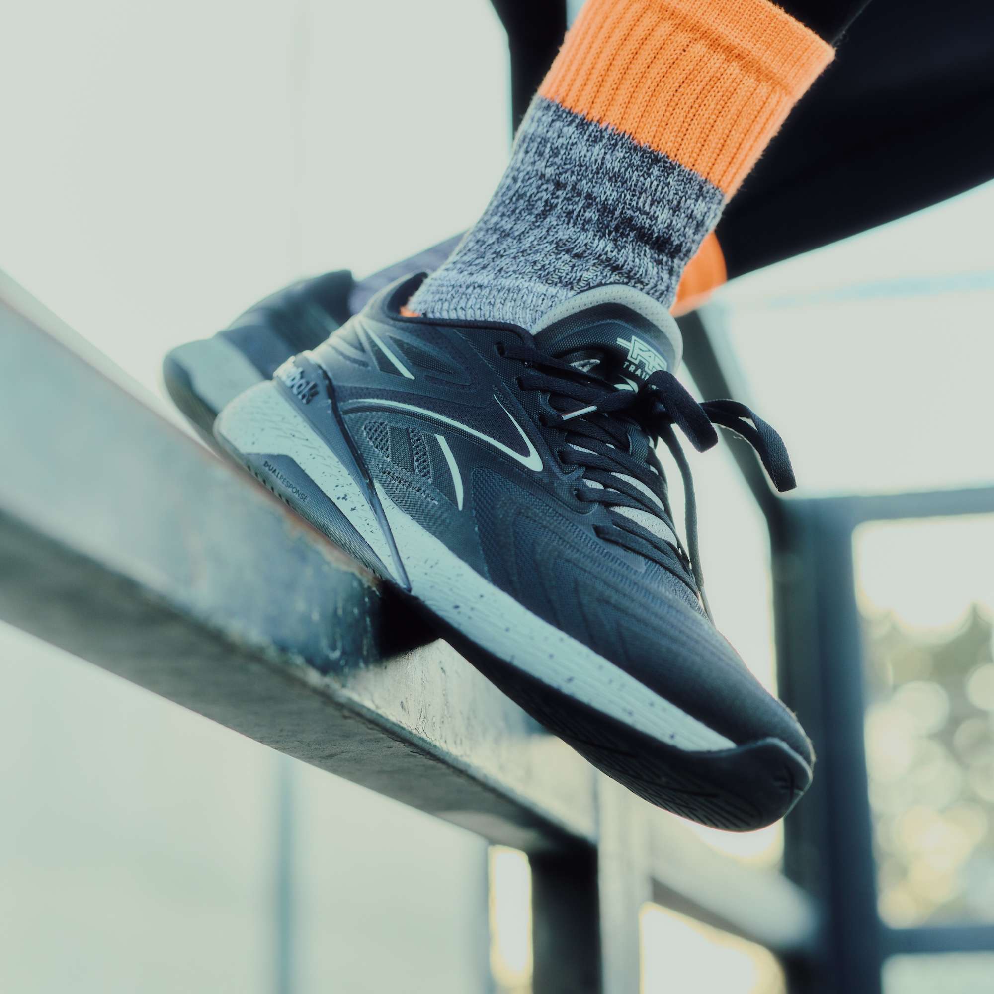 A close-up of a person’s foot in the Reebok x F45 Training Nano X5 Edge, standing on an outdoor metal rail. The focus is on the black performance workout shoe by Reebok, highlighted against a blurred background.