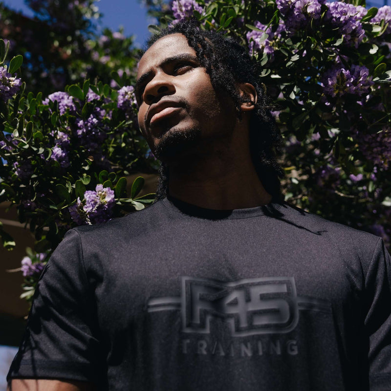 A man in a black Men's Reebok x F45 Training T-Shirt by Reebok stands outdoors before green leaves and purple flowers, gazing thoughtfully upward as sunlight illuminates his face.