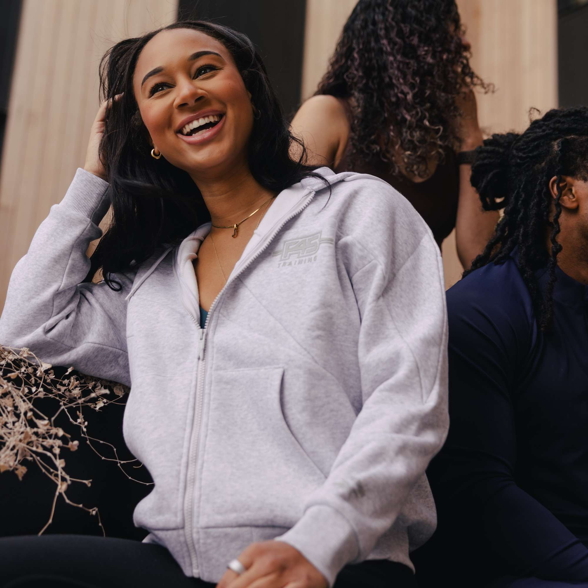 A smiling woman wears the Women's Reebok x F45 Training Relaxed Zip Hoodie as she sits outdoors on steps with two others, in front of a light-colored building.