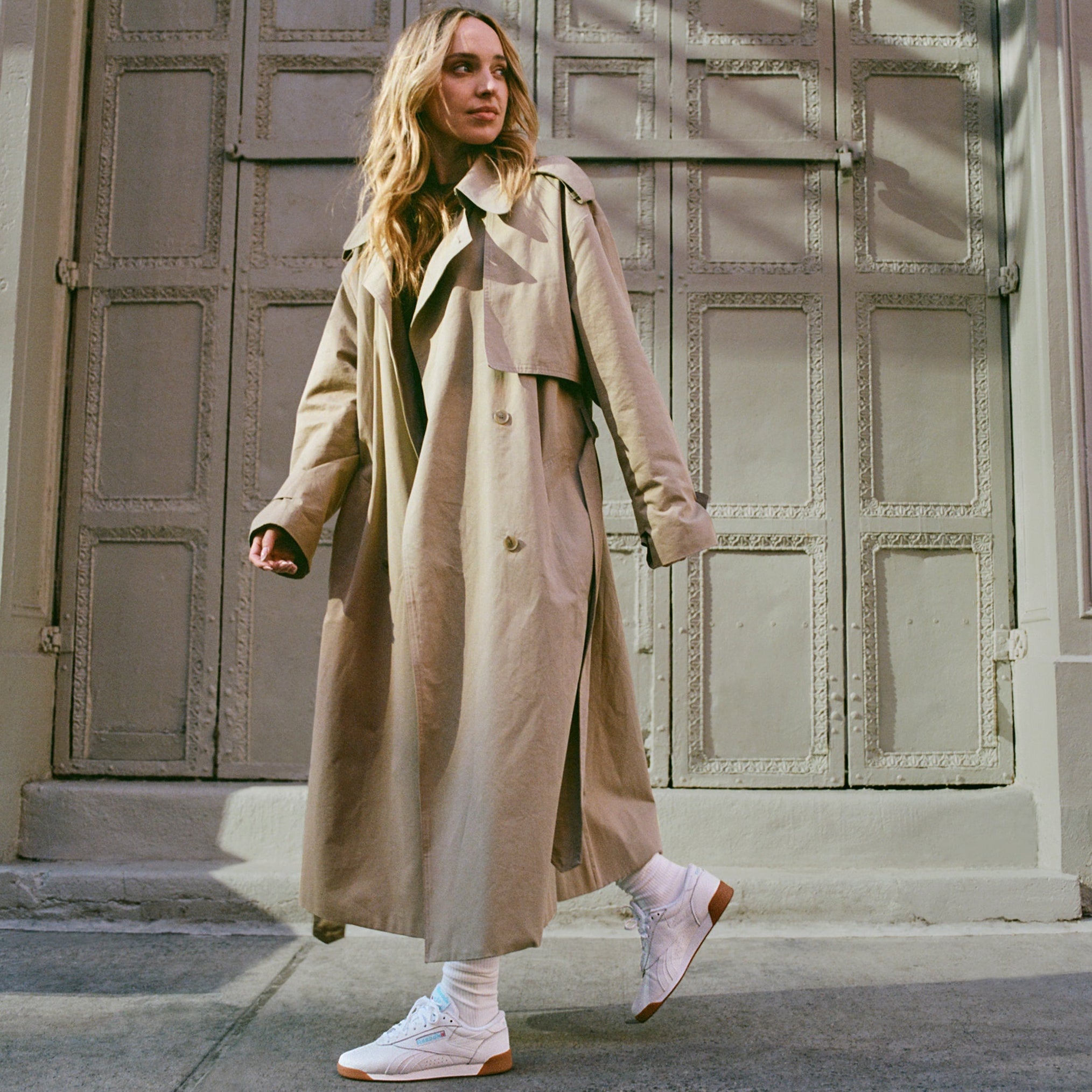 Woman in a beige trench coat walking on a street with a textured wall in the background