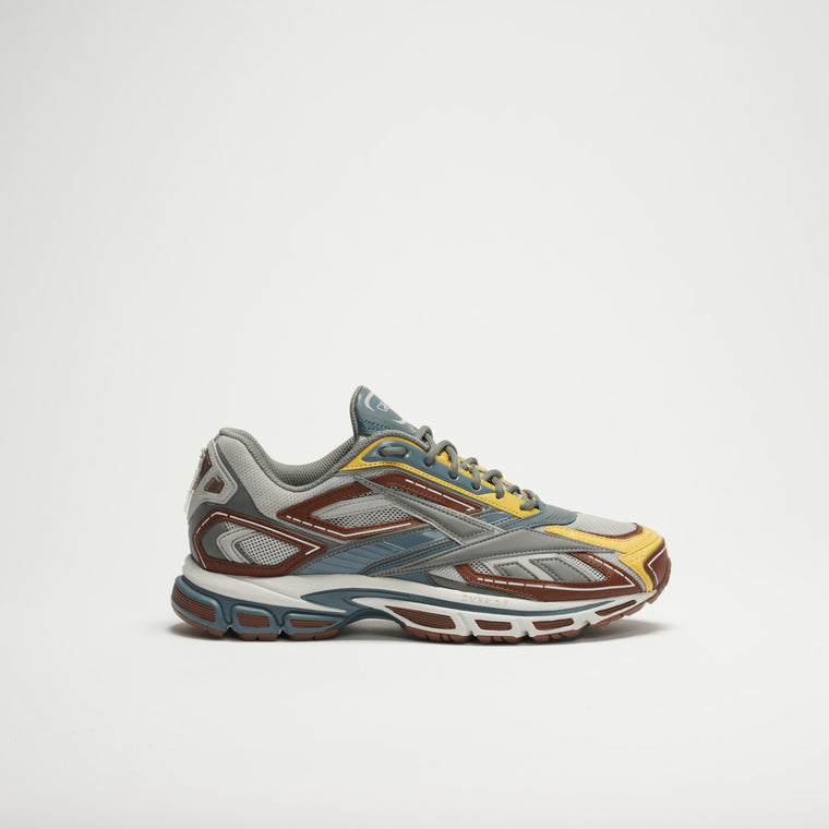 The Reebok Premier Road Ultra LTD Shoes showcase a modern, layered design in grey, blue, brown, and yellow shades with a dual-density midsole. Displayed from the side on a plain white background.