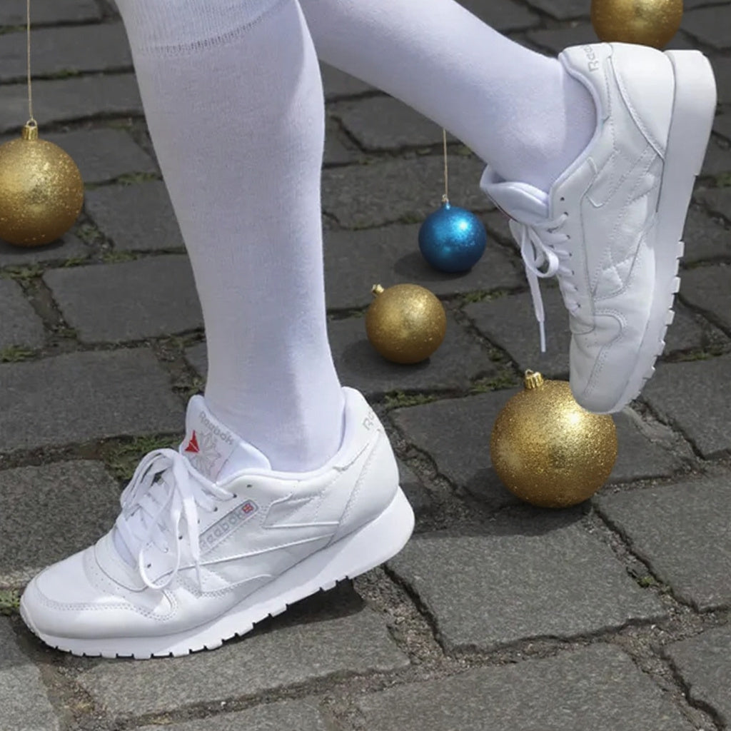 White leather sneakers worn with white tights, shown on a cobblestone surface with hanging gold and blue Christmas ornaments.