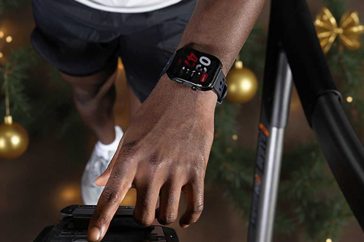 A close-up of a person's hand and wrist wearing a black smartwatch with a digital fitness display, standing near a treadmill in a decorated indoor setting with holiday lights and ornaments.
