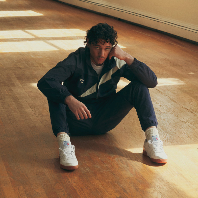 A man wears a dark-colored Men’s Classics Track Top with a white chevron detail, matching track pants, and white sneakers while sitting on a wooden floor.