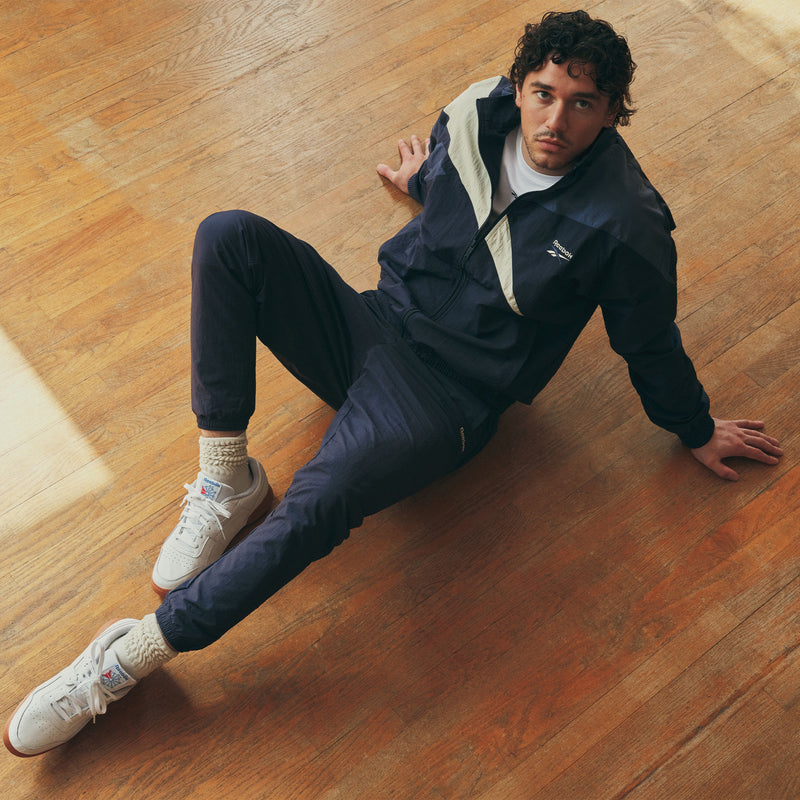 A man wearing a navy blue Reebok Men’s Classics Track Top with a white chest panel, matching track pants, and white sneakers with tan soles, sitting on a wooden floor.