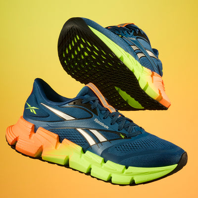 A pair of blue Reebok athletic shoes with neon green and orange soles are shown against a yellow gradient background. One shoe is upright, while the other is tilted to display the textured sole.