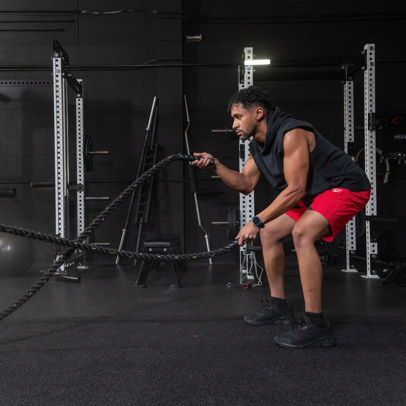 A man in a black sleeveless hoodie and red shorts trains with battle ropes in a gym, using the Reebok Pulse Smartwatch by Reebok to optimize his workout amid fitness equipment and racks.