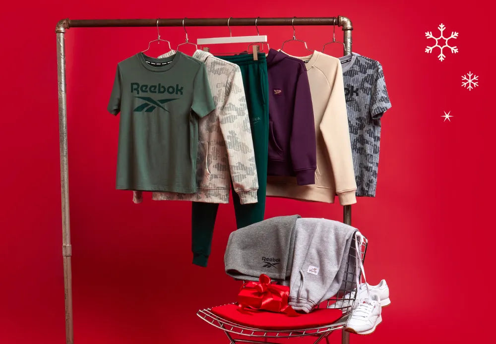 A clothing rack displays assorted Reebok athletic wear against a red background with snowflake graphics. A pair of sneakers and folded clothes with a red bow sit on a red chair below the rack.