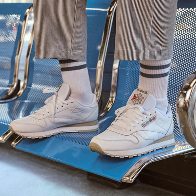 A person wearing white Reebok sneakers with white socks and striped trousers, sitting on a blue metal bench.