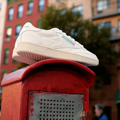 White Reebok sneaker on a red mailbox with blurred urban background