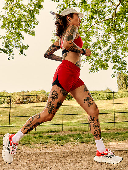 A tattooed female runner in a red sports bra and red athletic shorts sprints outdoors on a dirt path, wearing a white visor, white socks, and white running shoes with red accents.