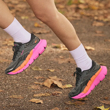 A person running outdoors on a leaf-covered path, wearing white socks and black running shoes with bright pink and orange soles.