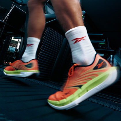 Close-up of a person running on a treadmill, wearing bright orange and green Reebok sneakers and white socks with a red Reebok logo. The image captures motion blur, emphasizing speed and movement.