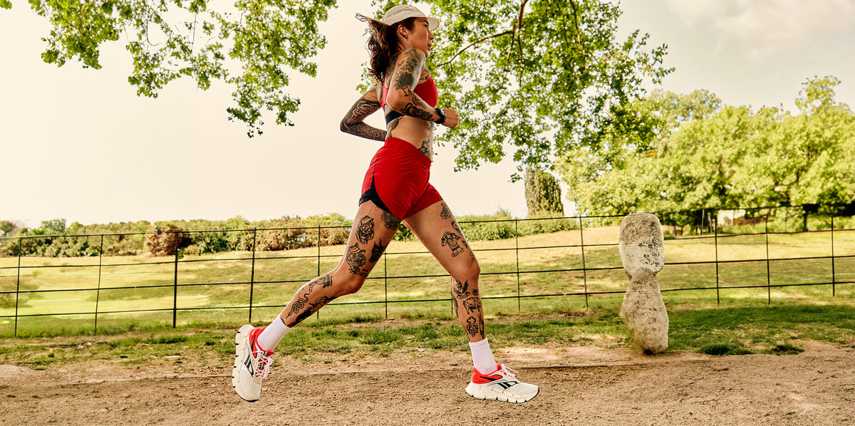 A woman running outdoors on a dirt path wearing a red athletic crop top, matching red shorts, white running socks, and white running shoes with red accents. She is captured mid-stride against a background of green trees and grass.
