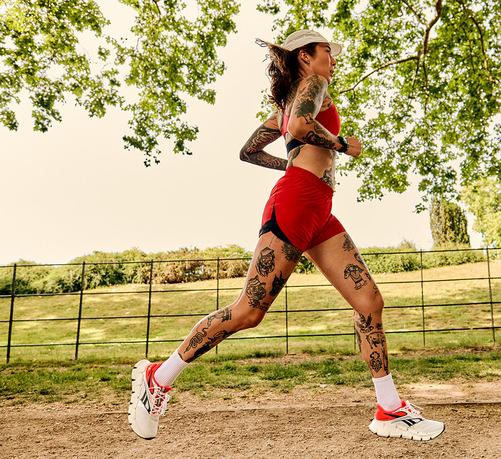 A woman with tattoos is running outdoors on a dirt path wearing a red sports bra, red athletic shorts, white running socks, and white running shoes with red accents. Trees and a fence are visible in the background.