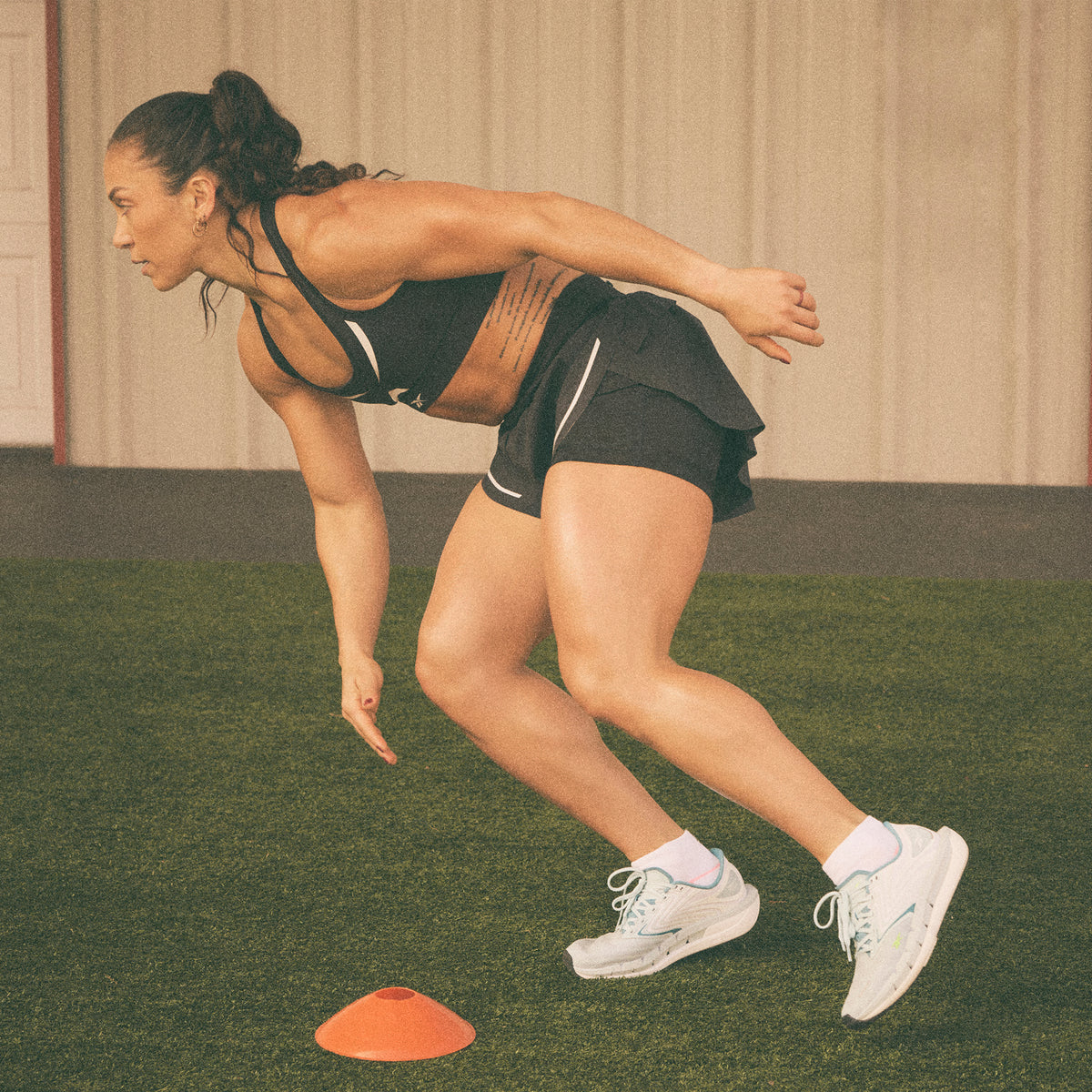 A woman in athletic wear crouches and leans forward with one arm extended, running a drill around an orange cone on artificial turf indoors.