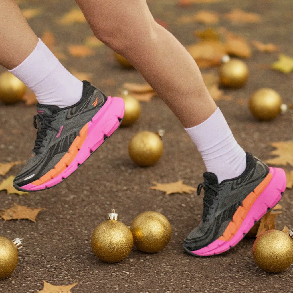 A person wearing gray and pink running shoes with orange accents, white socks, and bare legs, stepping onto a dark ground scattered with gold Christmas ornaments and fallen leaves.