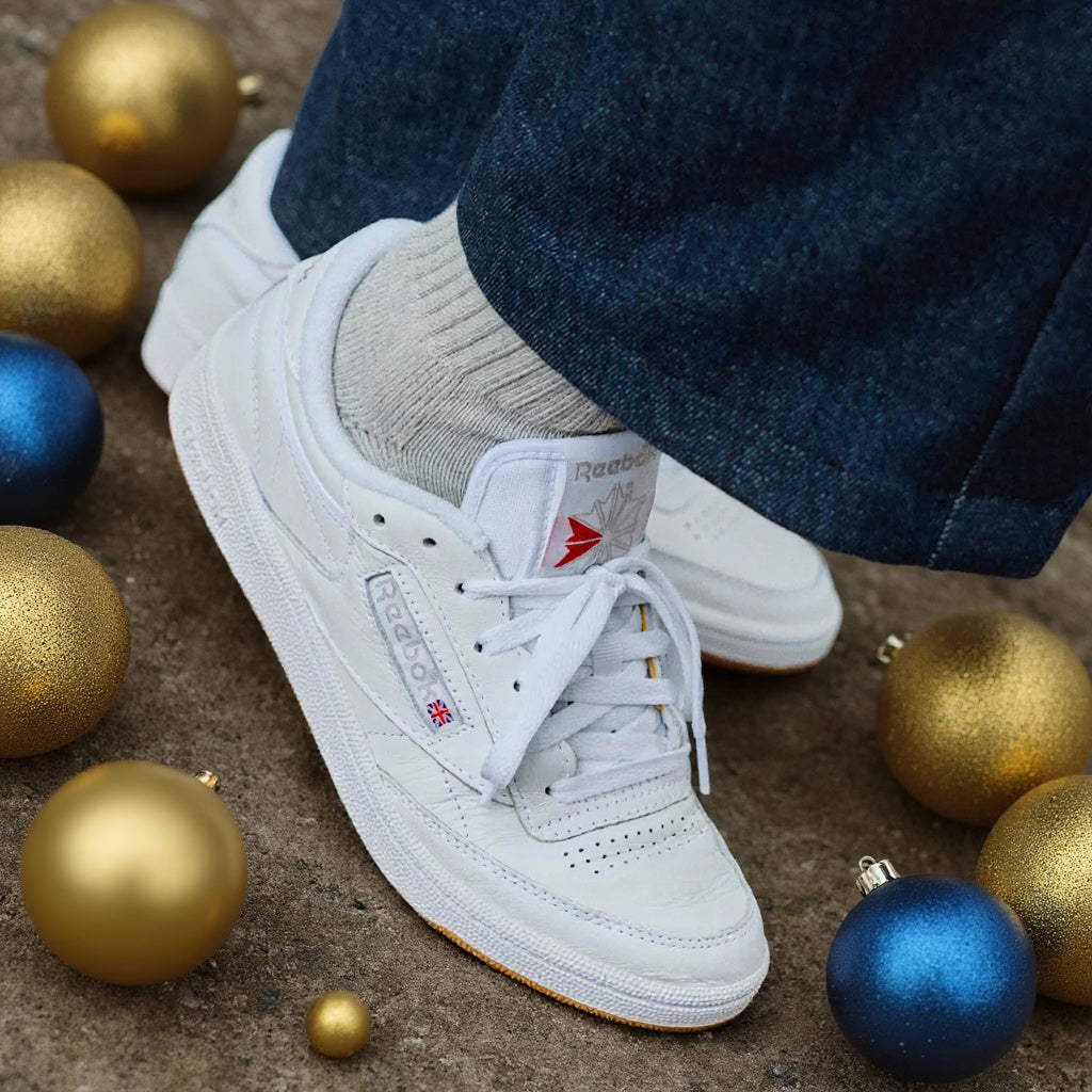 White Reebok Classic Leather sneakers worn with gray socks and blue jeans, surrounded by gold and blue Christmas ornaments on a concrete surface.