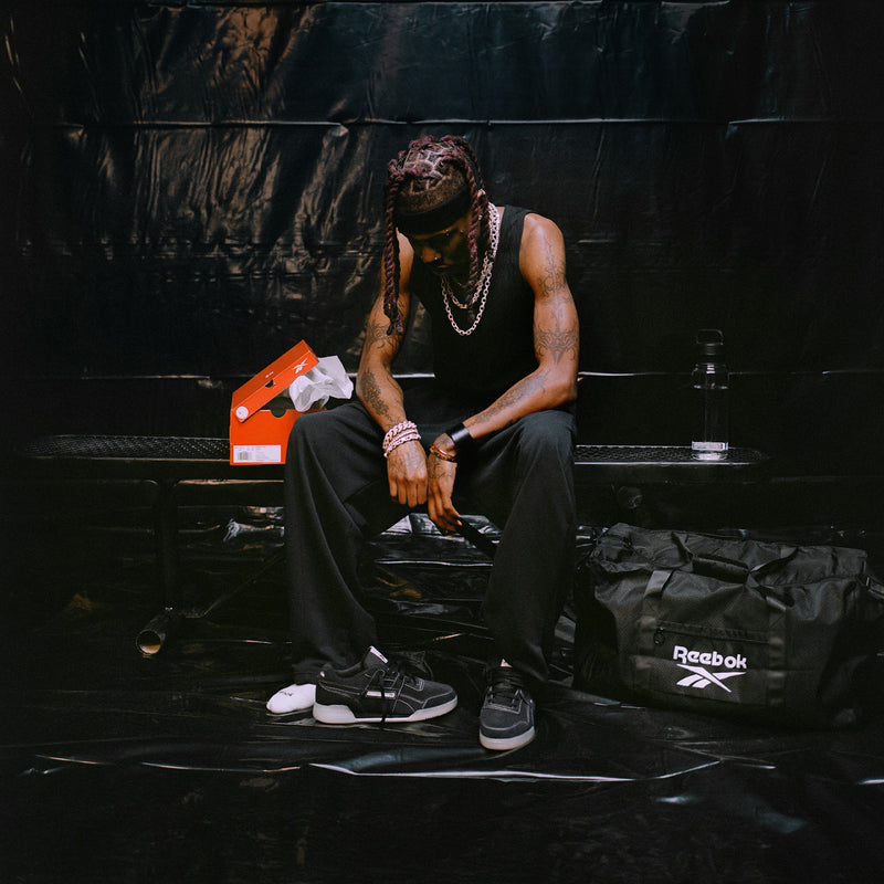A person dressed in dark athletic wear sits on a bench, gazing downward. Next to them, against a black plastic backdrop, are a shoebox, water bottle, and Reebok duffel bag—subtle nods to their commitment to fitness. Their outfit is elevated by the Reebok Workout Plus Shoes, which feature a sleek suede upper for timeless style that effortlessly fits their modern routine.