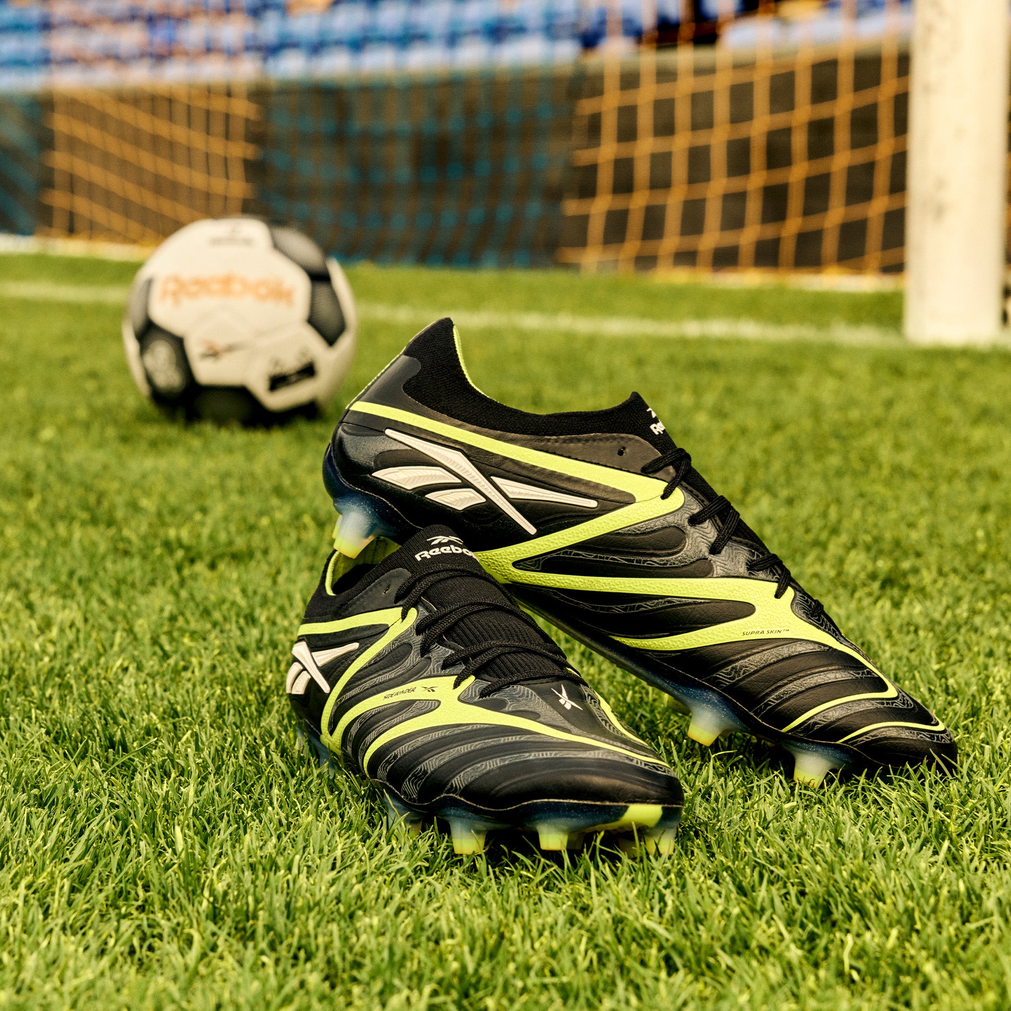Black and yellow soccer cleats on a grass field with a soccer ball and goal in the background.