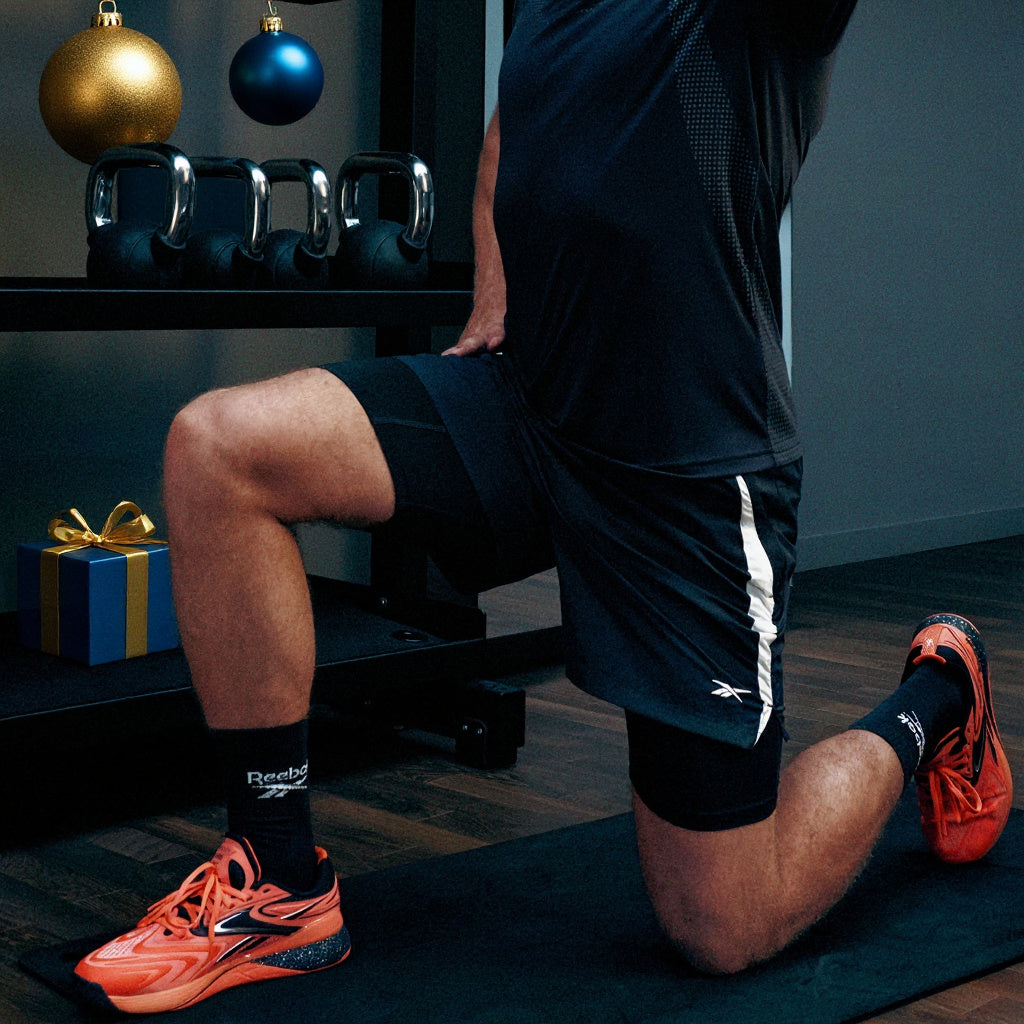 A person in a lunge position wearing black athletic shorts with a white side stripe, black Reebok socks, and bright orange athletic shoes on a black exercise mat in a gym setting.