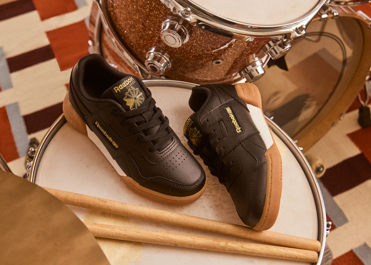 A pair of Reebok Recharge 2000 low-top sneakers in black leather with gold and white accents, styled on a drum kit with drumsticks and a patterned floor in the background.
