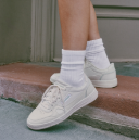 A person wearing white crew socks and white low-top sneakers, standing on a concrete step with a light-colored skirt visible at the top of the frame.