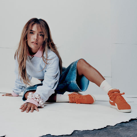 A woman sitting on the floor wearing orange Reebok Women’s Freestyle Hi Shoes with double ankle straps and white crew socks.