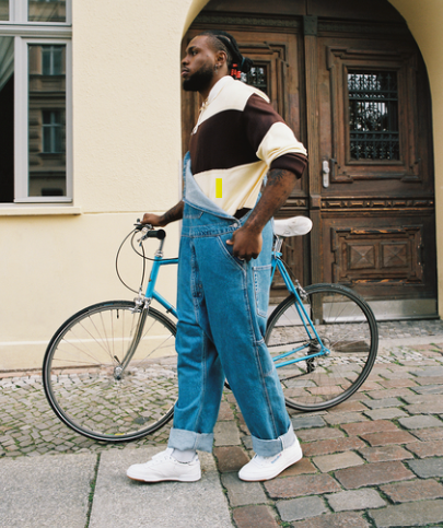 A person wearing blue denim overalls, a brown and white sweater, and white sneakers stands on a cobblestone street beside a blue bicycle, with wooden doors and a yellow building in the background.