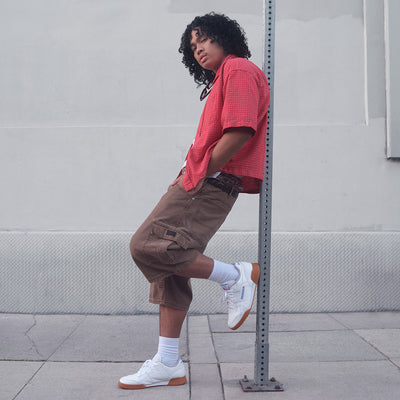 Person wearing a red shirt and brown cargo shorts leaning against a pole.