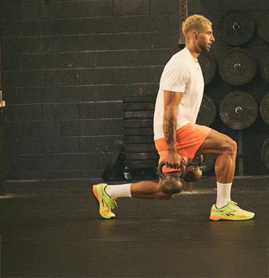 A man wearing a white shirt, orange shorts, and neon shoes performs a lunge while holding a kettlebell in each hand inside a gym with black walls and weight plates.