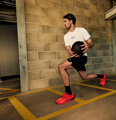 A man in athletic wear, including a white t-shirt, black shorts, and bright red Adidas sneakers, holds a black medicine ball while performing a lunge in an indoor concrete parking structure.