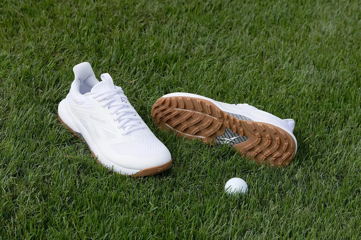 A pair of white athletic shoes with brown soles are placed on green grass next to a white golf ball.