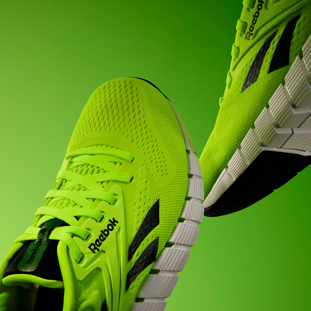Close-up of Reebok Men's Nano Gym Training Shoes in neon green with white soles, black logo accents, and a performance foam collar, displayed against a gradient green background.