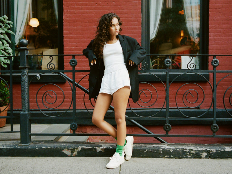 A young woman with curly hair stands on a sidewalk in front of a red brick building, wearing a black jacket, white tank top, white shorts, green socks, and white sneakers. She leans on a railing and looks to the side.