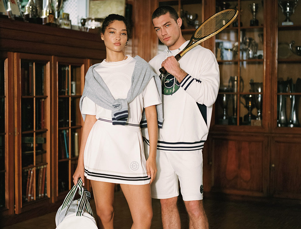 Two people in tennis attire standing in a room with wooden shelves.