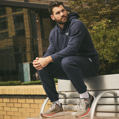 A man with a beard sits on a silver bench outdoors, wearing a navy blue hoodie, matching sweatpants, white socks, and gray sneakers. He looks to the side, with greenery and a building in the background.
