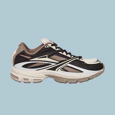 The Reebok Premier Road Modern Shoes feature a beige, white, black, and dark brown color scheme with a chunky sole and layered futuristic design, shown on a light blue background.
