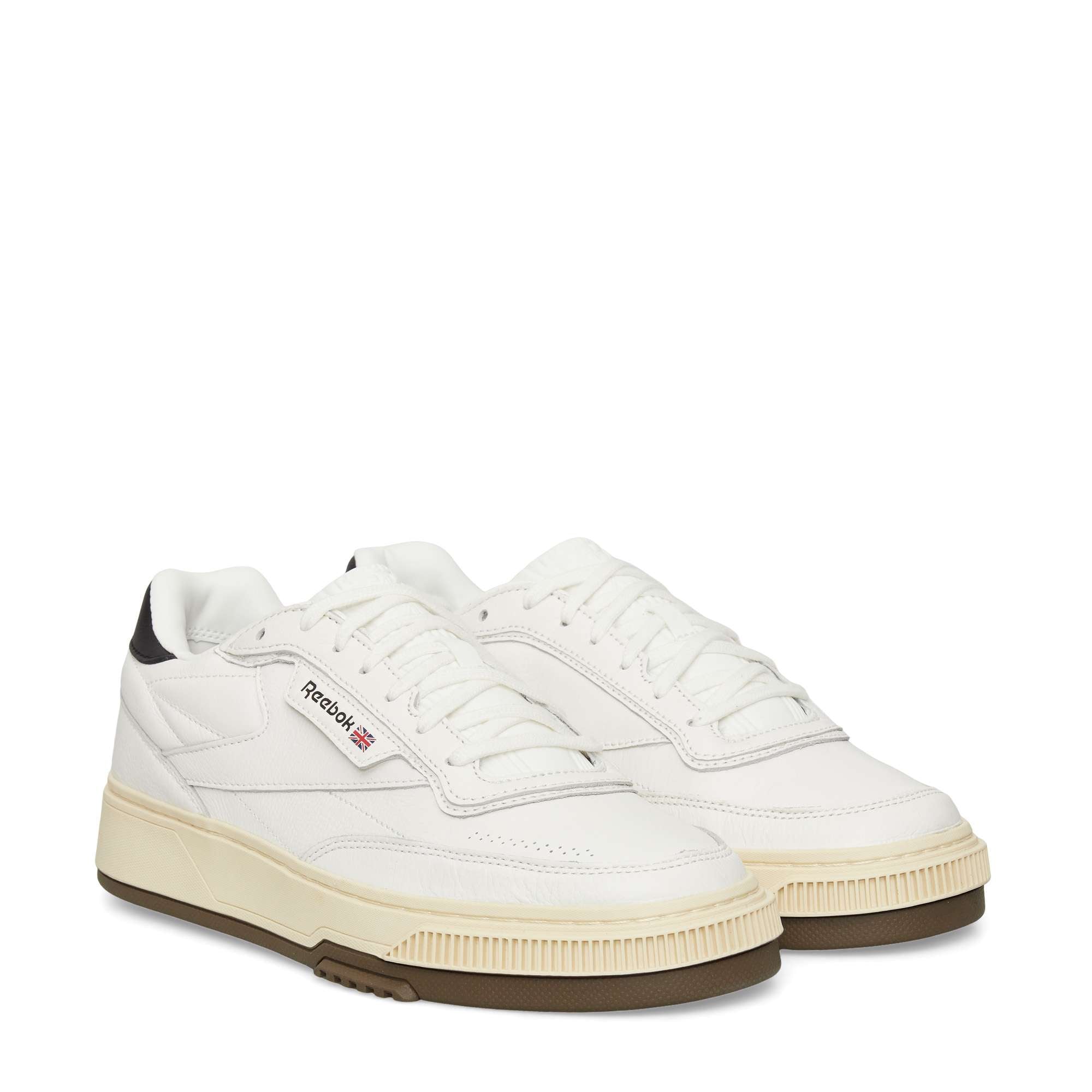 A pair of white Reebok sneakers with black detailing on the heel, off-white midsoles, gum rubber outsoles, and the Reebok logo on the side. The shoes have a retro design with lace-up fronts.