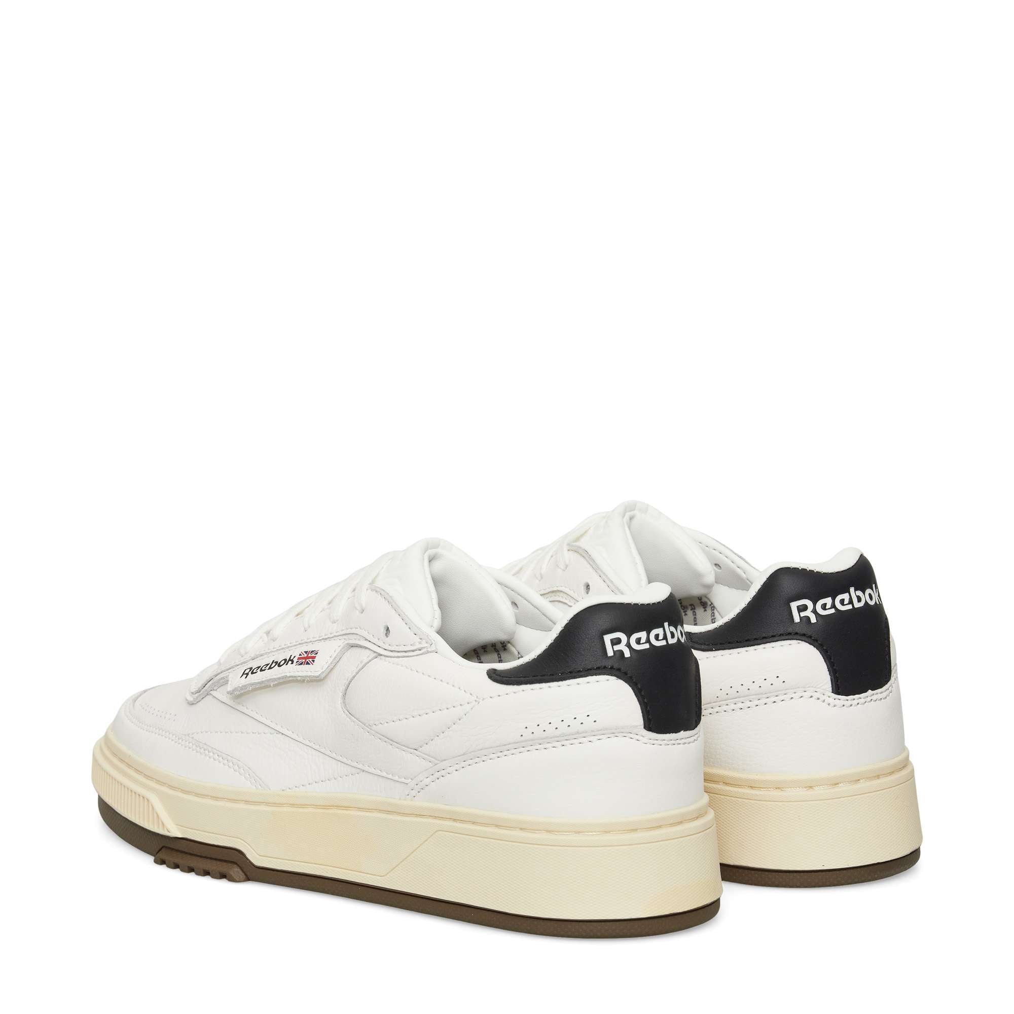 A pair of white Reebok sneakers with black heel tabs, gum-colored outsoles, and the Reebok logo visible on the side and back. The shoes are positioned at an angle, showing both the side and back.