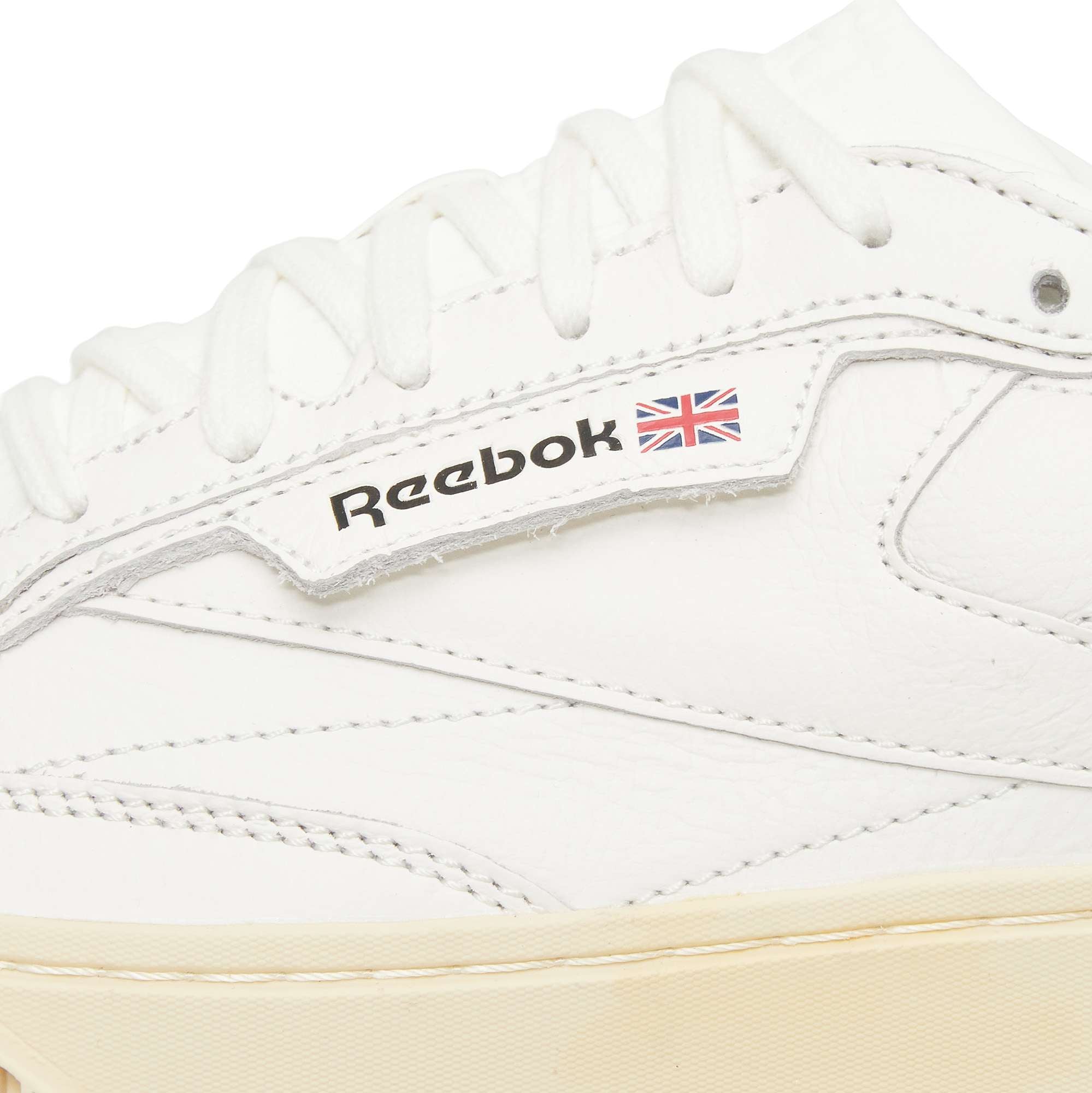 Close-up of a white Reebok sneaker, showing white laces, stitching, and the Reebok logo with a small Union Jack flag on the side. The sole is a light beige color.
